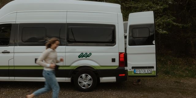 Ein Grand California Van steht auf einem Waldstück. Eine Frau ist verschwommen zu sehen, sie läuft am Camper vorbei und lacht.